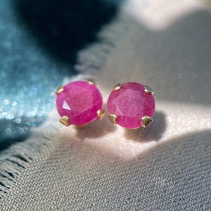 May include: A pair of ruby stud earrings. Each earring features a round, faceted, bright pink gemstone set in a gold-colored metal. The earrings are displayed on a light-colored, textured fabric.