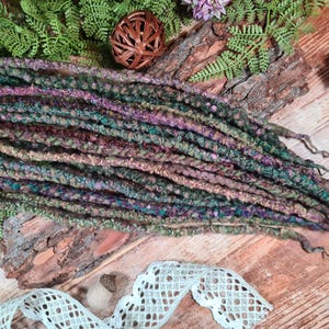 Op de afbeelding: Handgemaakte dreadlocks in groen, paars en bruin, gepresenteerd op een houten oppervlak met decoratieve accenten. De getextureerde dreadlocks, waarschijnlijk gemaakt van garen, creëren een bohemien, natuurlijke esthetiek. De opstelling omvat varens en een decoratieve houten bal.