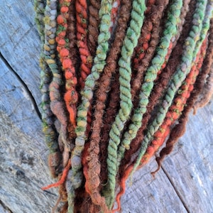 Puede incluir: Primer plano de un conjunto de rastas coloridas. Las rastas son una mezcla de naranja, verde, marrón y gris. La textura de las rastas es gruesa y retorcida, con algunos mechones sueltos en los extremos. Las rastas están dispuestas en un patrón en cascada.