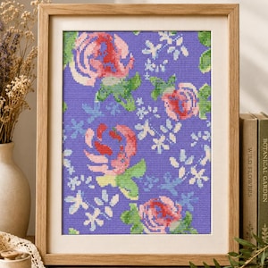 Könnte beinhalten: Ein gerahmtes Kreuzstich-Blumenmuster mit rosa und roten Rosen, grünen Blättern und weißen Akzenten auf lila Hintergrund. Das Kunstwerk befindet sich in einem Holzrahmen.