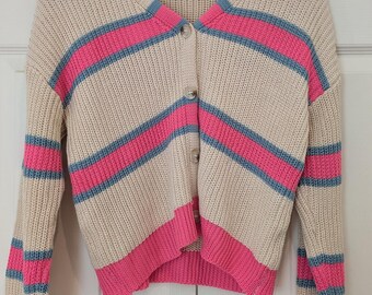 Può includere: Cardigan beige con bottoni e righe orizzontali rosa e blu. Il maglione ha maniche lunghe e scollo a V. I polsini e l'orlo inferiore sono rosa a tinta unita. Il cardigan è appeso a una gruccia di legno.
