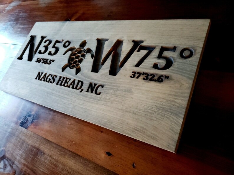 May include: A wooden sign with the coordinates N35&deg; 56'55.5" W75&deg; 37'32.6" and the text "NAGS HEAD, NC" carved into it. The sign also features a carved sea turtle.