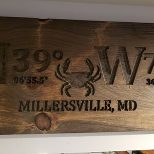 May include: A dark brown wooden sign with the coordinates N39&deg; 06'55.5" W76&deg; 36'39.8" and the text "MILLERSVILLE, MD" carved into the wood. A small crab is carved in the centre of the sign.