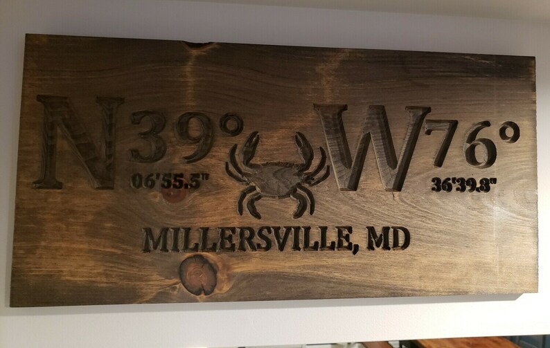May include: A dark brown wooden sign with the coordinates N39&deg; 06'55.5" W76&deg; 36'39.8" and the text "MILLERSVILLE, MD" carved into the wood. A small crab is carved in the centre of the sign.