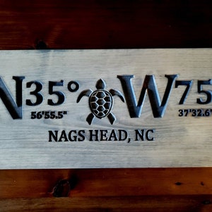 May include: A wooden sign with the coordinates N35&deg; 56'55.5" W75&deg; 37'32.6" and the text "NAGS HEAD, NC" carved into it. A sea turtle is carved in the centre of the sign.