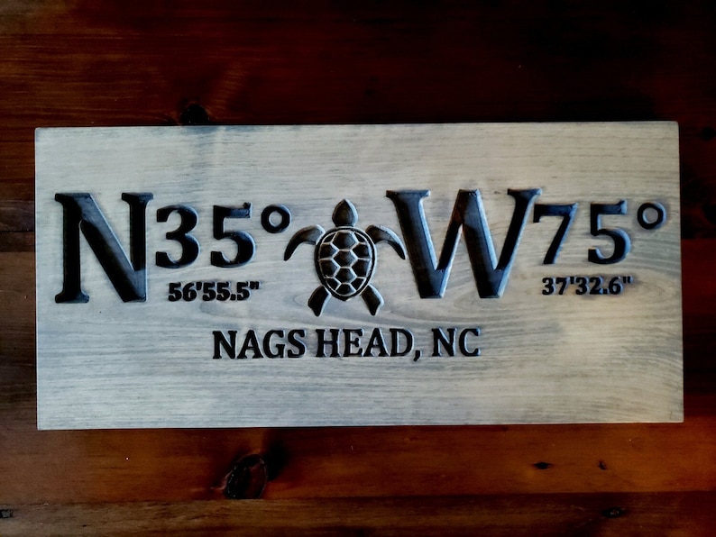 May include: A wooden sign with the coordinates N35&deg; 56'55.5" W75&deg; 37'32.6" and the text "NAGS HEAD, NC" carved into it. A sea turtle is carved in the centre of the sign.