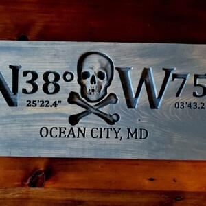 May include: A wooden sign with a carved skull and crossbones design. The sign reads "N38&deg; 25'22.4" W75&deg; 03'43.2" OCEAN CITY, MD".
