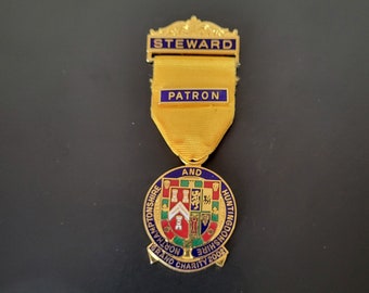 Puede incluir: Medalla dorada con un emblema azul y rojo. La medalla lleva las palabras "STEWARD" y "PATRON" en una cinta dorada. El emblema dice "NORTHAMPTONSHIRE AND HUNTINGDONSHIRE GRAND CHARITY 2002".