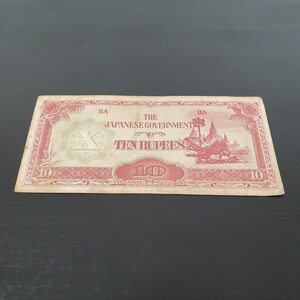 Puede incluir: Un billete vintage de diez rupias del Gobierno japonés. El billete presenta tinta roja y verde con texto e ilustraciones. El billete rectangular se muestra sobre una superficie oscura.