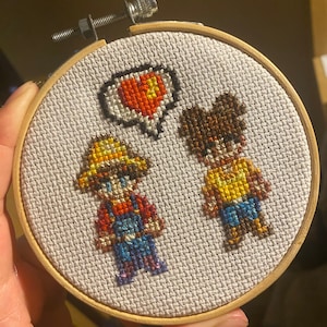 May include: Cross-stitch embroidery hoop with two figures, a person wearing a yellow hat and overalls and another person wearing a yellow shirt and blue jeans. A speech bubble with a red heart is between the figures.