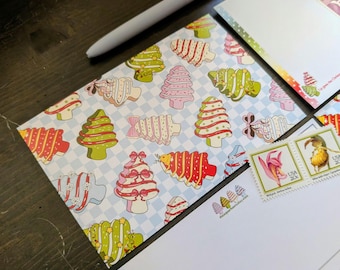 May include: A collection of Christmas-themed stationery, including patterned envelopes and postcards. The envelopes feature a repeating pattern of colorful Christmas trees on a light blue checkered background. Postage stamps and a pen are also visible.