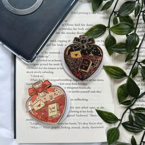 Peut inclure: Deux épingles en émail en forme de cœur. L'une représente un flacon avec les mots "With Love" et une enveloppe. L'autre représente un flacon avec les mots "Till Death" et un livre. Les deux épingles ont des accents rouges et dorés.