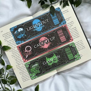 May include: Three decorative bookmarks with ticket designs, each featuring a different color scheme and text: "LIGHTS OUT," "CAUGHT UP," and "GAME ON." The bookmarks are placed on an open book.
