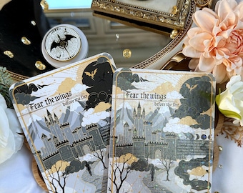 May include: Two rectangular art prints featuring a castle illustration with the text 'Fear the wings before the fire'. The design includes a stone path, bare trees, a dark sky with a dragon silhouette, and gold accents. A circular pin with a matching dragon design is visible in the background.