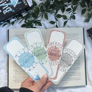 Puede incluir: Cuatro marcadores coloridos con temas elementales: Agua, Tierra, Fuego y Aire. Cada marcador presenta un diseño circular con símbolos astrológicos y el nombre del elemento. Los marcadores se sostienen sobre un libro abierto, con un libro y vegetación en el fondo.