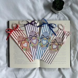 May include: Four decorative bookmarks with striped patterns and initial designs. Each bookmark features a different colour ribbon: red, purple, blue, and pink. The bookmarks are placed on an open book, with a small plant in the background.