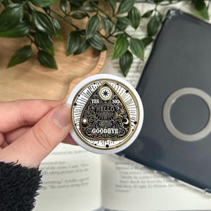 Pode incluir: Uma planchette redonda branca e dourada com as palavras "YES", "NO", "HELLO" e "GOODBYE" em preto. A planchette tem uma borda dourada e um desenho de estrela. A planchette é segurada em frente a um livro e uma capa cinza escuro.