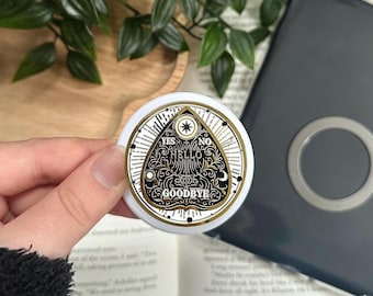 May include: A white and gold round planchette with the words "YES", "NO", "HELLO", and "GOODBYE" in black. The planchette has a gold border and a star design. The planchette is held in front of a book and a dark gray case.