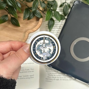 Op de afbeelding: Een ronde witte popsocket met een gouden rand en een zwaardontwerp. Het ontwerp toont een zwaard, wolken en sterren in blauw- en grijstinten. De popsocket wordt in een hand gehouden, met een boek en een telefoonhoesje op de achtergrond.