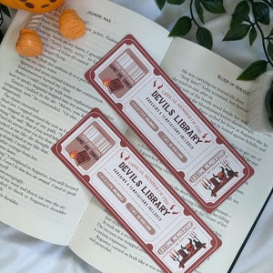 Puede incluir: Tres marcapáginas de color burdeos con el texto "OFFICAL MEMBER OF THE DEVILS LIBRARY" y "LIFETIME MEMBERSHIP". Cada marcapáginas tiene un diseño de boleto con un código de barras y una ilustración de diablo. Los marcapáginas están sobre un libro abierto.