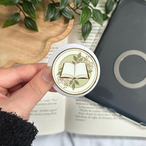 May include: A white and gold pop socket with an open book design, pink flowers, and green leaves. The pop socket is held in front of an open book and a grey electronic device.