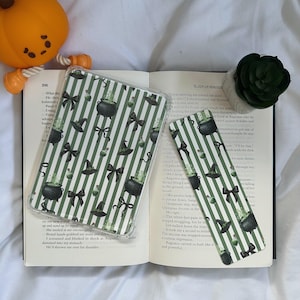 May include: A clear phone case and bookmark with a green and white striped pattern. The design includes black witch hats, bows, and cauldrons with cacti. The items are displayed on an open book with a pumpkin and succulent.