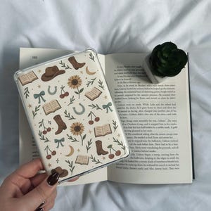 以下が含まれることがあります： クリーム色の背景にカウボーイブーツ、本、ヒマワリ、サクランボ、蹄鉄、リボンが繰り返されるパターンが特徴の透明な携帯電話ケース。ケースは開いた本の上に置かれています。