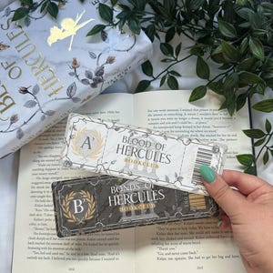 Könnte beinhalten: Ein Buch mit dem Titel "Blood of Hercules" und zwei Lesezeichen. Die Lesezeichen tragen die Aufschrift "Blood of Hercules Bookclub" und "Bonds of Hercules Bookclub". Das Buch hat ein Blumendesign und eine goldene Fee.