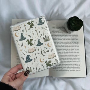 Op de afbeelding: Een witte telefoonhoes met een fantasierijk patroon van groene draken, tovenaarshoeden, sterren, manen, zwaarden, bogen en pijlen, en open boeken. De hoes ligt op een open boek.