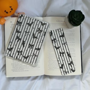May include: A clear phone case and a bookmark with a black and white striped pattern featuring bats and bows. The items are placed on an open book with text. A small orange pumpkin decoration is visible.