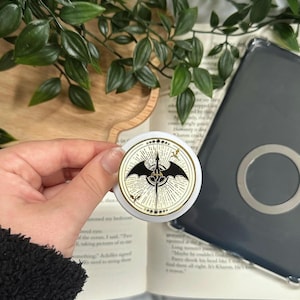 Puede incluir: Un accesorio circular blanco y dorado con un diseño negro en forma de murciélago, brújula y detalles de estrellas. Se sostiene sobre un libro abierto y una funda de teléfono.