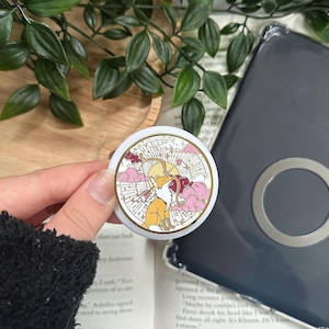 Puede incluir: Un soporte circular blanco para teléfono con un diseño de zorro de dibujos animados, con un sol y nubes rosas. El zorro es naranja y blanco, sobre un fondo dorado y crema. El soporte se sostiene sobre un libro abierto.