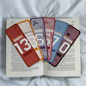 May include: Six colorful bookmarks with different designs and numbers. The bookmarks are made of cardstock and feature a variety of illustrations, including a hockey stick, a crown, a heart, and a football. The numbers on the bookmarks are 3, 3, 5, 7, and 0.