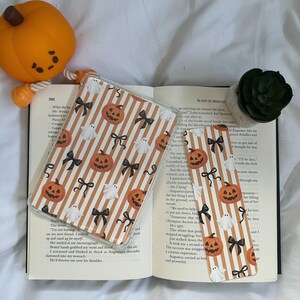 Puede incluir: Funda de teléfono transparente y marcapáginas con un diseño de Halloween. La funda y el marcapáginas muestran calabazas naranjas, fantasmas blancos y lazos negros sobre un fondo a rayas. También se ve un pequeño juguete de calabaza naranja.