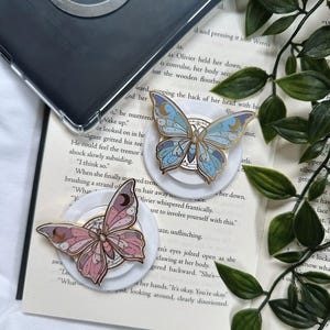 May include: Two butterfly-shaped phone grips in the image. One is light blue with gold accents, and the other is pink with a crescent moon design. Both are on white circular bases, set against a book page and greenery.