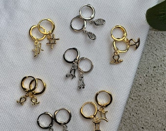 May include: A set of 10 gold and silver hoop earrings with zodiac symbols. The earrings are arranged on a white surface. The symbols include Gemini, Libra, Sagittarius, Pisces, Cancer, Virgo, Capricorn, Aquarius, Aries, and Taurus.