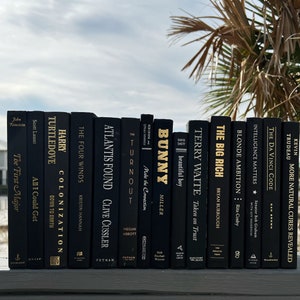 May include: A collection of black books with gold lettering on the spines. The books are arranged in a row on a wooden surface. The titles of the books include "The Four Winds", "Atlantis Found", "Turnout", "Bunny", "Terry Waite", "The Big Rich", "Blonde Ambition", "Intelligence Matters", "The Da Vinci Code", and "More Natural Cures Revealed".