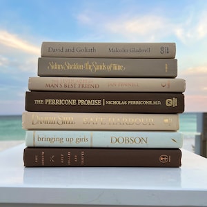Könnte beinhalten: Ein Stapel von acht Büchern mit braunen, beigefarbenen und weißen Einbänden. Die Bücher sind von groß nach klein angeordnet. Die Titel der Bücher sind sichtbar, darunter "David und Goliath", "Sidney Sheldon - Die Sanduhr der Zeit", "Die sieben Lebensalter des besten Freundes des Menschen", "Das Perricone-Versprechen", "Danielle Steel Safe Harbour", "Mädchen erziehen" und "Paul Morrom Only Killers and Thieves".