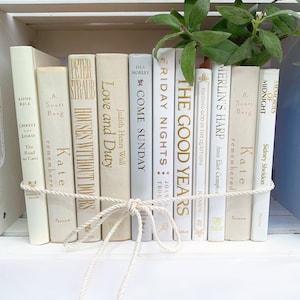 May include: A shelf of white books with gold lettering, tied together with a white rope. The titles include "Love and Duty", "Come Sunday", "Friday Nights", "The Good Years", "Merlin's Harp", and "Memories of Midnight".