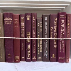 Puede incluir: Una colección de libros de tapa dura en tonos burdeos y marrón, dispuestos en una estantería blanca. Los títulos incluyen "Virgins of Paradise", "Bordeaux" y "Night Shall Overtake Us". Una cuerda blanca asegura los libros, creando una exhibición decorativa.