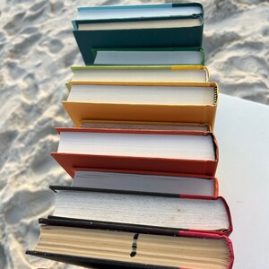 May include: A stack of eight books with different colored covers. The books are arranged in a vertical stack, with the top book being a light blue color and the bottom book being a dark red color.