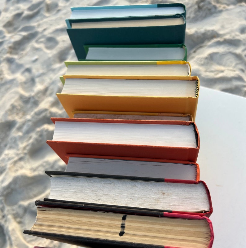 May include: A stack of eight books with different colored covers. The books are arranged in a vertical stack, with the top book being a light blue color and the bottom book being a dark red color.