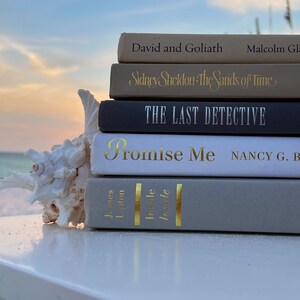 May include: A stack of five books on a white table with a seashell in the foreground. The books are titled "David and Goliath", "Sidney Sheldon - The Sands of Time", "The Last Detective", "Promise Me", and "Inside/Inside".
