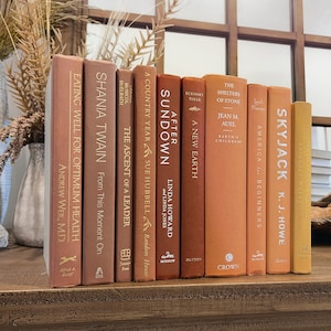 May include: A collection of ten books with orange, brown, and yellow covers. The books are arranged vertically on a wooden shelf. The titles of the books include "Eating Well for Optimum Health", "Shania Twain", "The Ascent of a Leader", "A Country Year", "After Sundown", "A New Earth", "The Shelters of Stone", "Earth's Children", "America for Beginners", and "Skyjack".