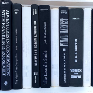 Könnte beinhalten: Ein Bücherregal mit einer Sammlung schwarzer und weißer Bücher. Die Titel der Bücher sind sichtbar, darunter "Adventures in Conservation with Franklin D. Roosevelt", "The Ultimate Weapon is No Weapon", "The Lizard's Smile", "Final Harvest", "Honor and Blood", "Every Spy a Prince".