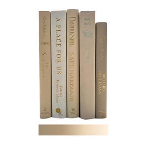 May include: A collection of five hardcover books in varying shades of beige and cream. The spines display titles and author names in gold lettering. The books are arranged vertically, suggesting a decorative display or a curated bookshelf.