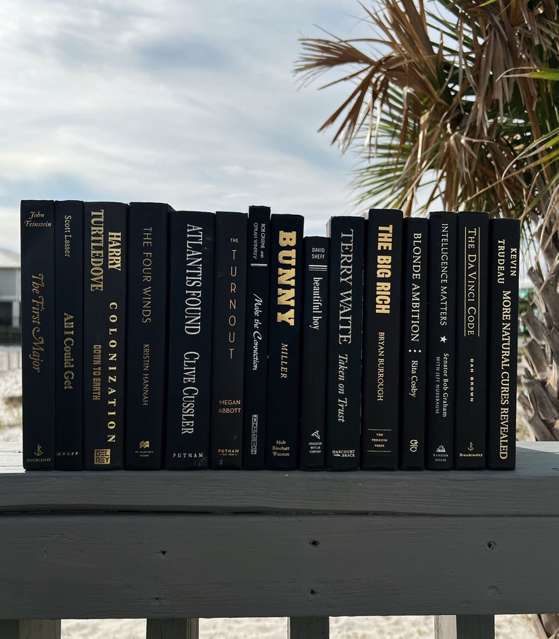 May include: A collection of black books with gold lettering on the spines. The books are arranged in a row on a wooden surface. The titles of the books include "The Four Winds", "Atlantis Found", "Turnout", "Bunny", "Terry Waite", "The Big Rich", "Blonde Ambition", "Intelligence Matters", "The Da Vinci Code", and "More Natural Cures Revealed".