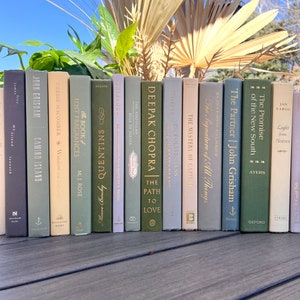 Könnte beinhalten: Eine Reihe dekorativer Bücher in verschiedenen Farben, von Salbeigrün bis Hellbeige. Titel wie "The Path to Love" und "Camino Island" sind sichtbar. Die Bücher sind auf einer Holzoberfläche angeordnet.