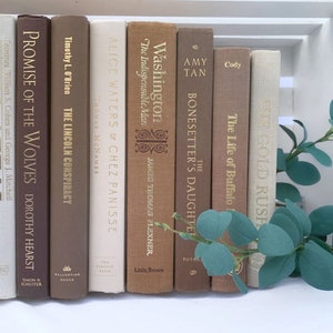 May include: A collection of vintage hardcover books with brown, beige, and white spines. The books are arranged in a row on a white surface. The titles of the books include "Promise of the Wolves", "The Lincoln Conspiracy", "Alice Waters & Chez Panisse", "Washington The Indispensable Man", "The Bonesetter's Daughter", "The Life of Buffalo", and "White Gold Rush".