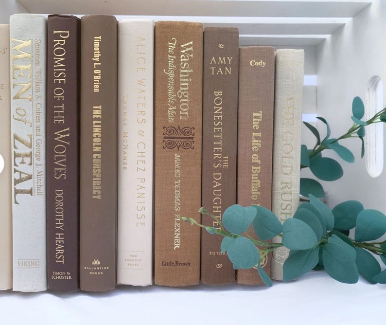 May include: A collection of vintage hardcover books with brown, beige, and white spines. The books are arranged in a row on a white surface. The titles of the books include "Promise of the Wolves", "The Lincoln Conspiracy", "Alice Waters & Chez Panisse", "Washington The Indispensable Man", "The Bonesetter's Daughter", "The Life of Buffalo", and "White Gold Rush".